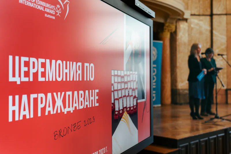 Екран с надпис „Церемония по награждаване“ и водещи на сцената